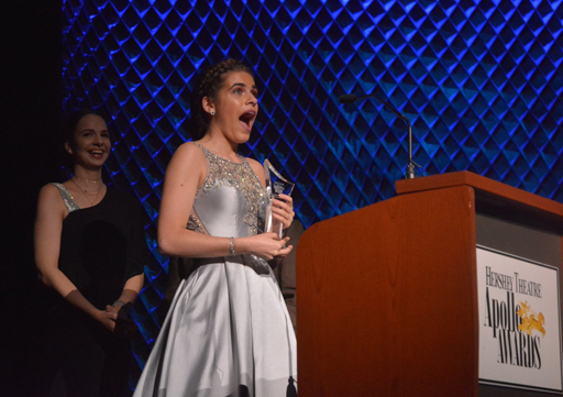 girl looking shocked yet excited after recieving an award on stage while wearing a beautiful silver gown