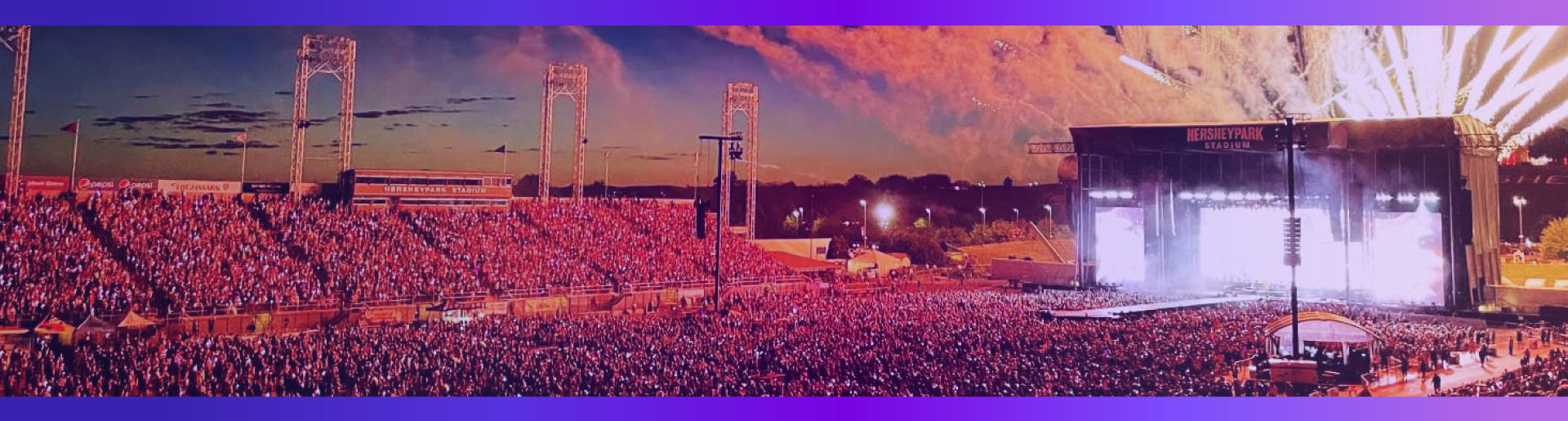 A high-angle view of a huge concert crowd at nighttime at Hersheypark Stadium, with the stage lit up and fireworks in the sky.